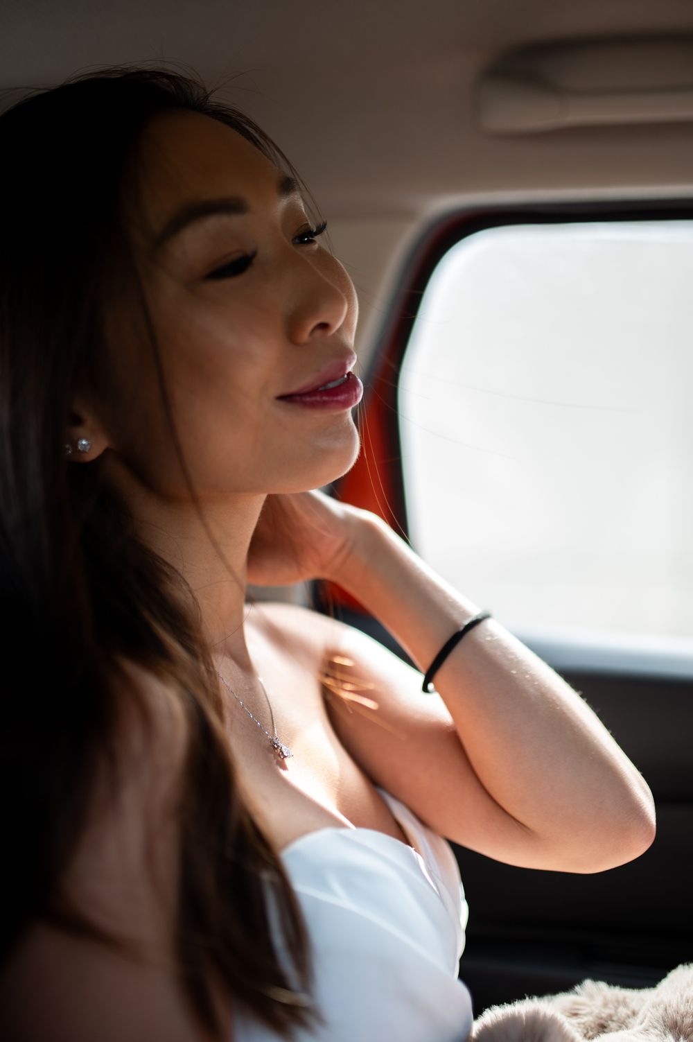 Young Asian woman in white top sitting in car backseat, wearing necklace and bracelet, posing gracefully in sunlight.