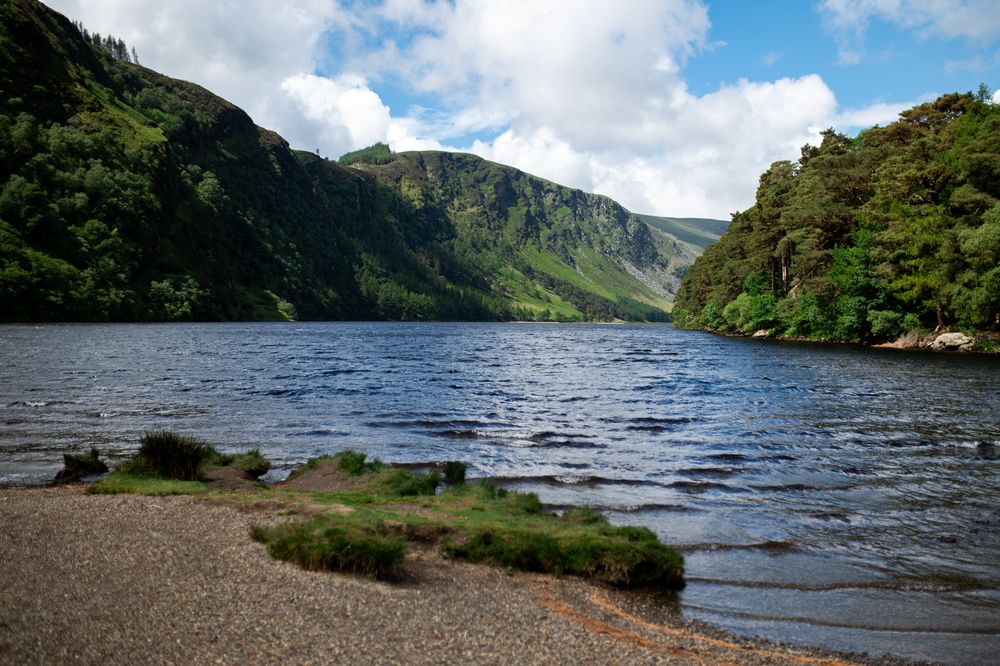 Scenic lake surrounded by lush green mountains and trees under a partly cloudy sky in Glendalough, Ireland.