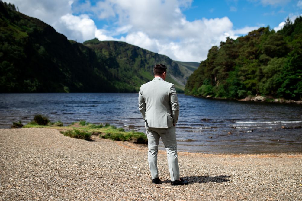 Groom in light grey suit standing alone gazing at a scenic mountain lake.