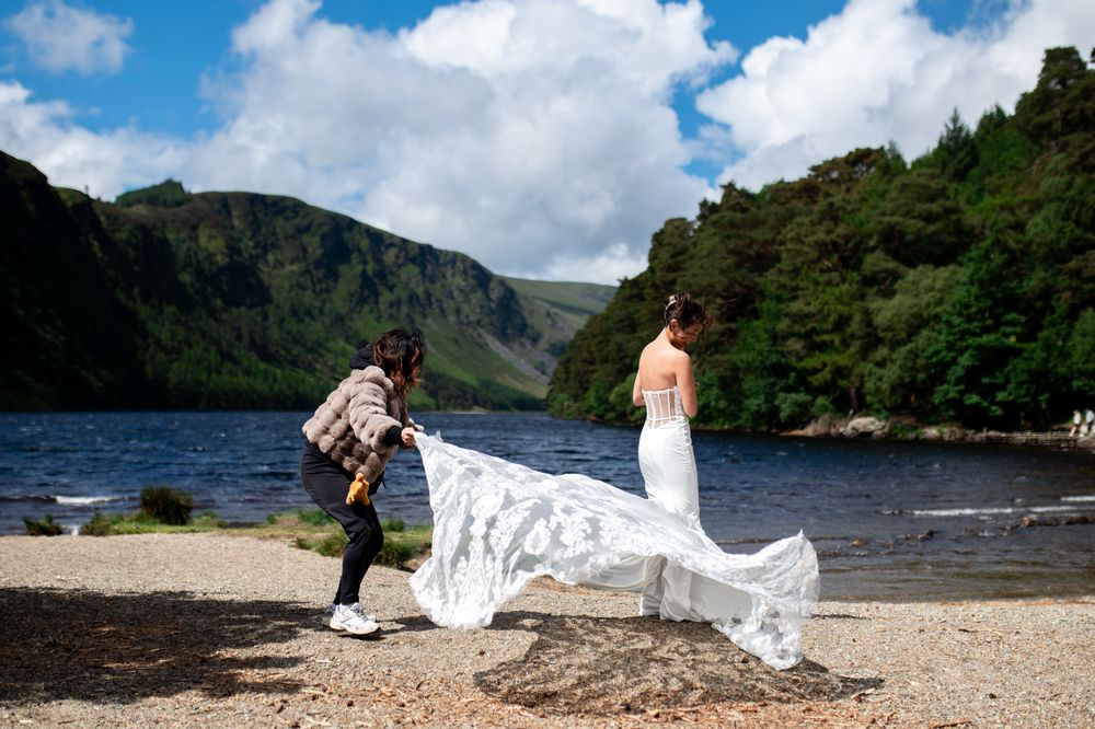 Photographer captures bride's lace wedding gown train on rocky lakeside with lush green mountains backdrop.