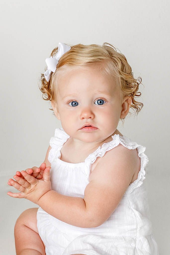 Adorable baby girl with blue eyes and curly blonde hair wearing a white dress and bow hair clip.