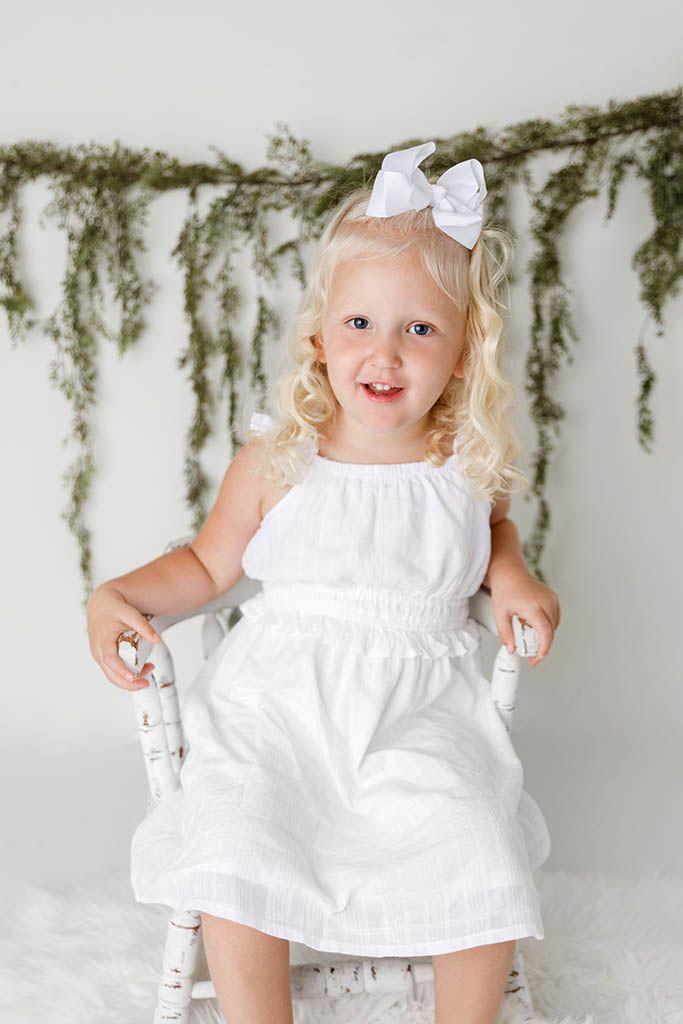 Smiling blonde toddler girl in white dress and bow sits on white chair with greenery backdrop.