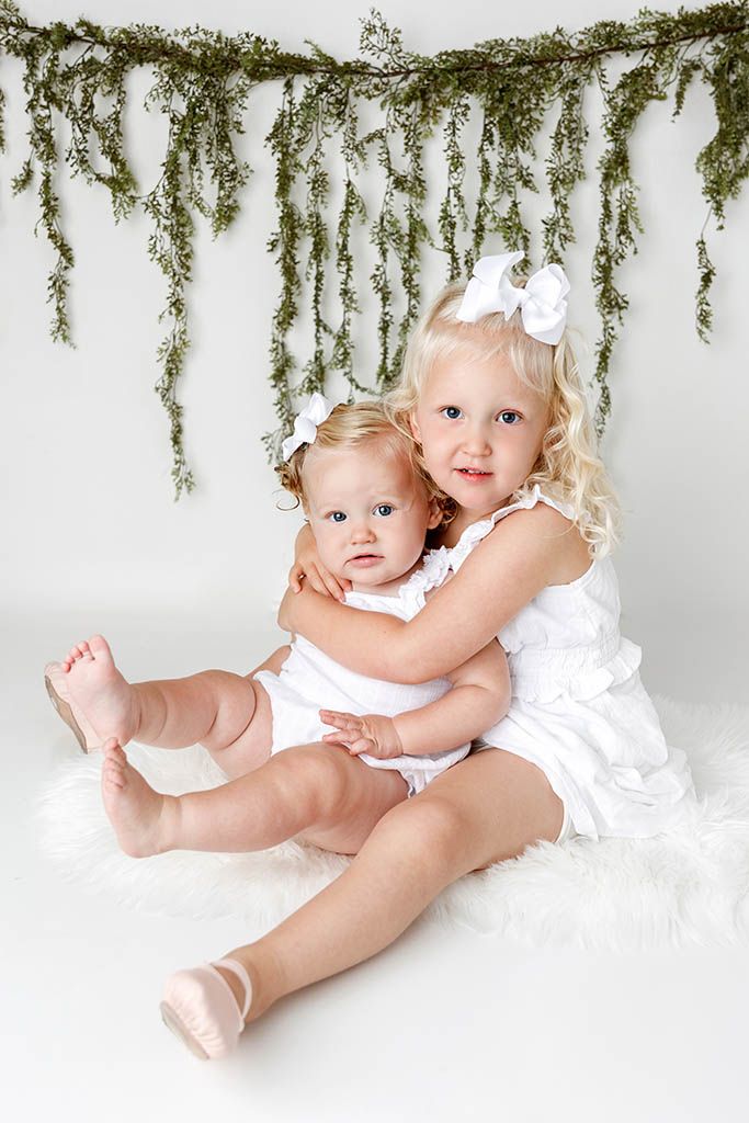 Two young sisters in white dresses pose together under hanging greenery for a portrait.