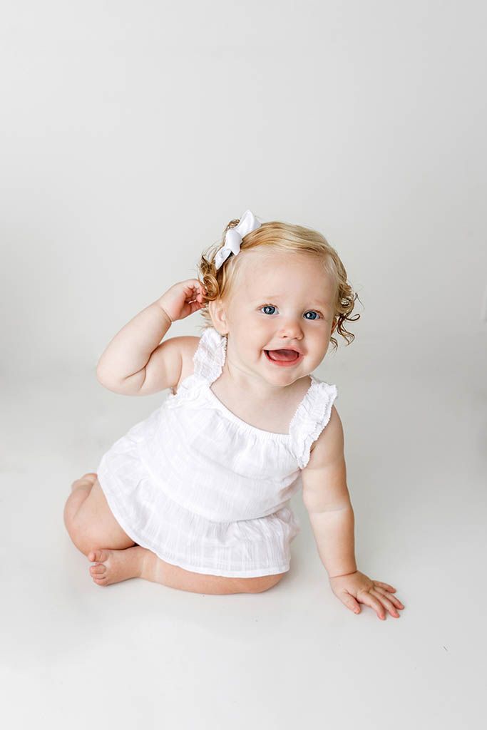 Smiling baby girl in white romper and bow sits on white floor during professional photo session.