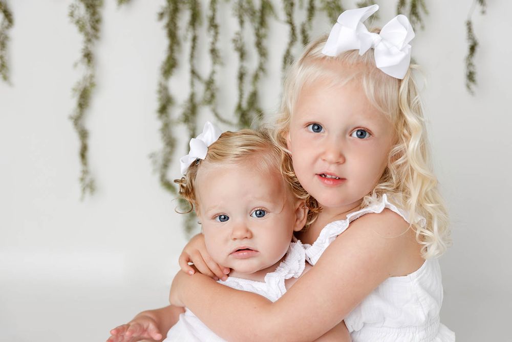 Two young blonde sisters in white dresses pose together, older girl hugging baby sibling with white bows in their hair.