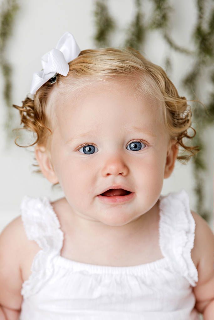 Close-up portrait of blue-eyed baby girl with curly blonde hair wearing white bow and white dress.