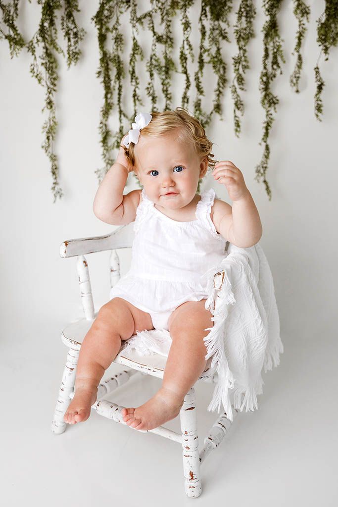 Baby girl in white romper sits on rustic white chair with hanging greenery background.