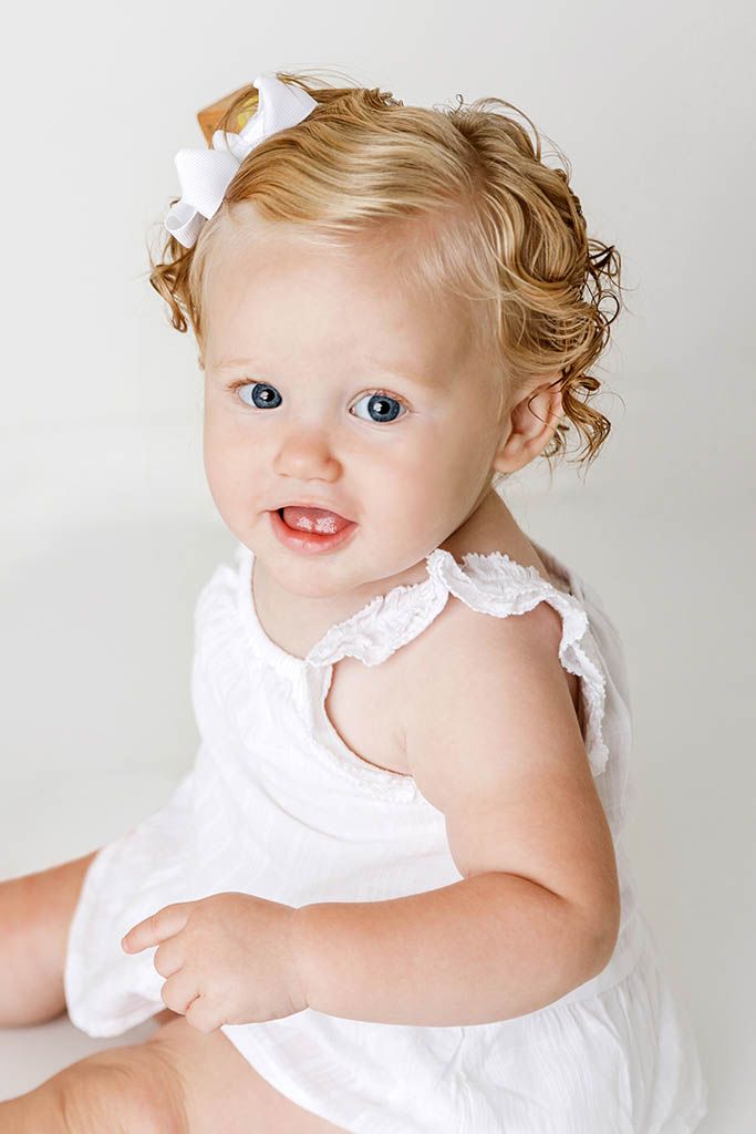 Adorable baby girl with curly blonde hair and blue eyes smiles in white lace romper and bow.