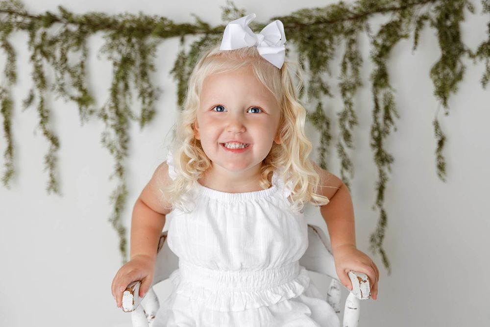 Happy toddler girl with blonde curls in white dress and bow smiles on chair with greenery backdrop.