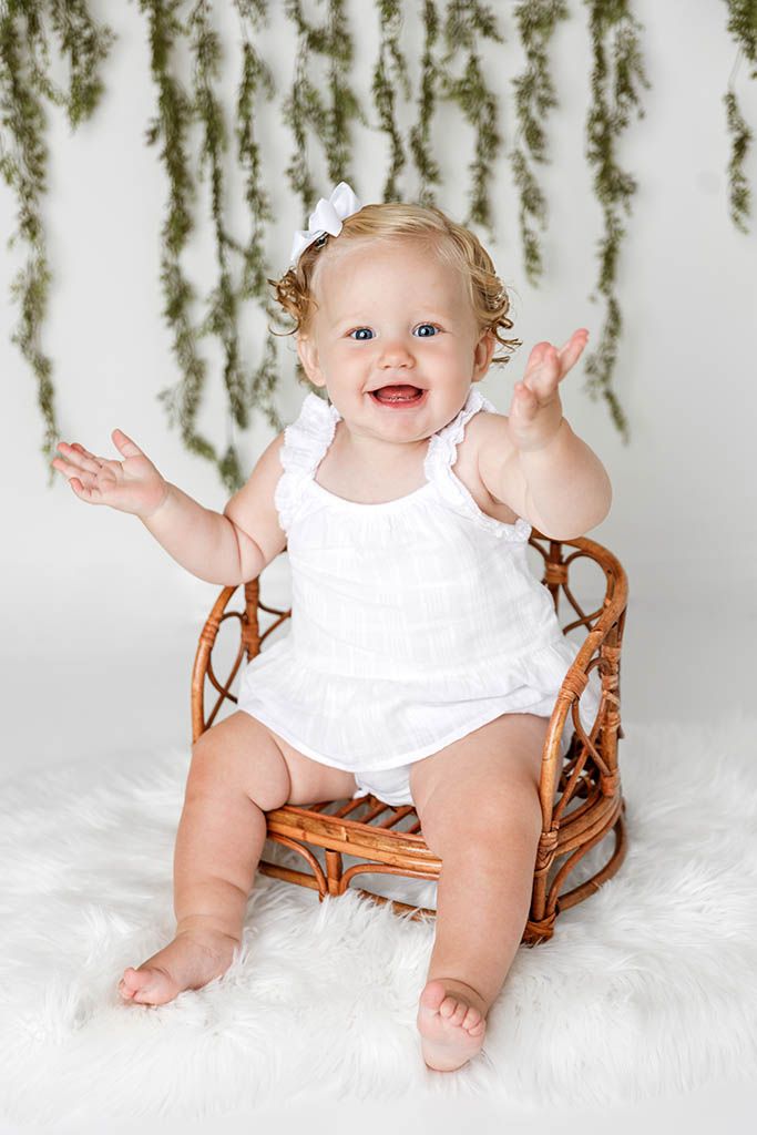Joyful baby girl in white romper claps hands while sitting in wicker chair with greenery backdrop.