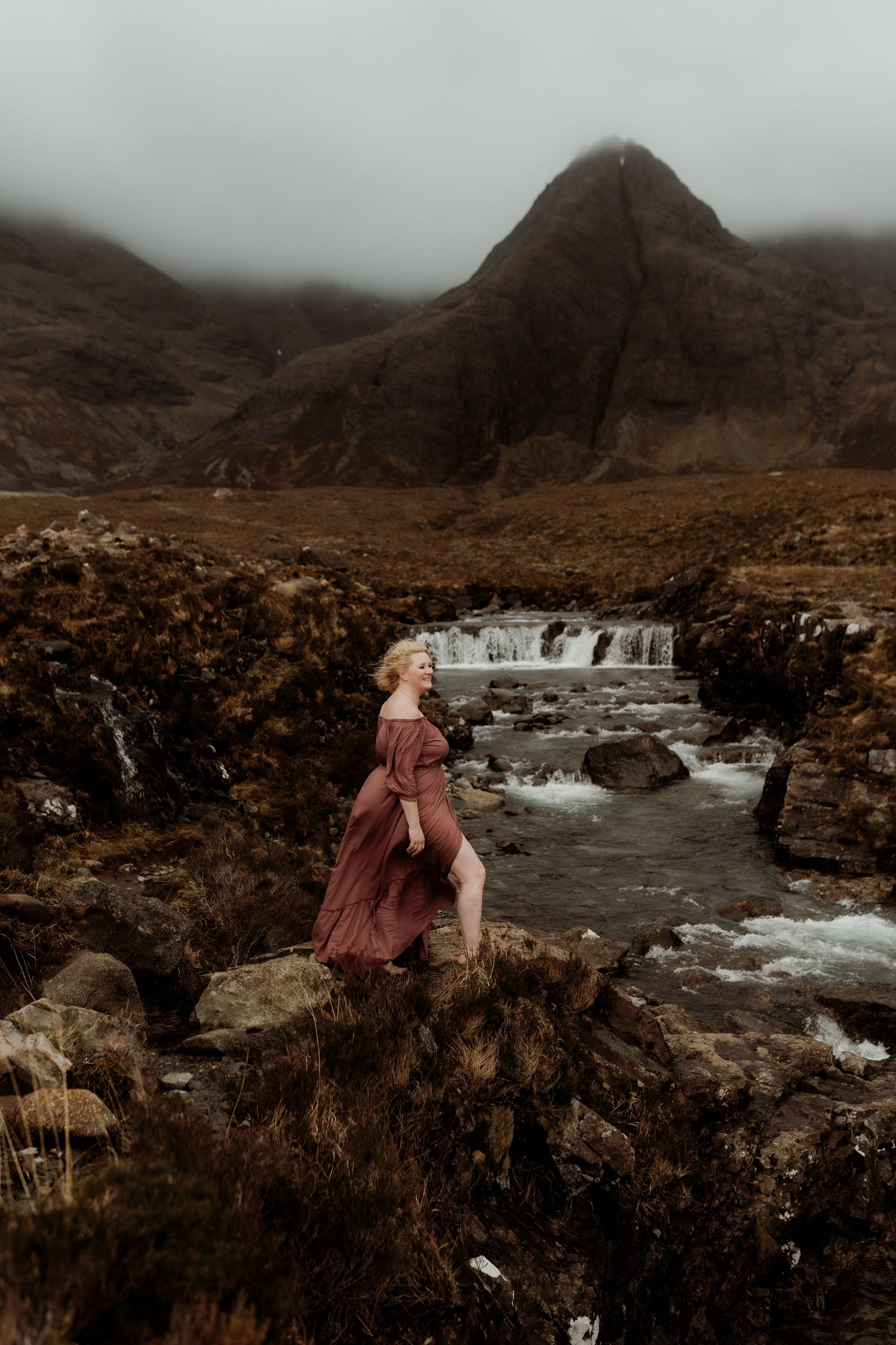 Woman in flowing burgundy dress standing on rocks by a misty mountain stream in a dramatic Scottish Highland landscape.