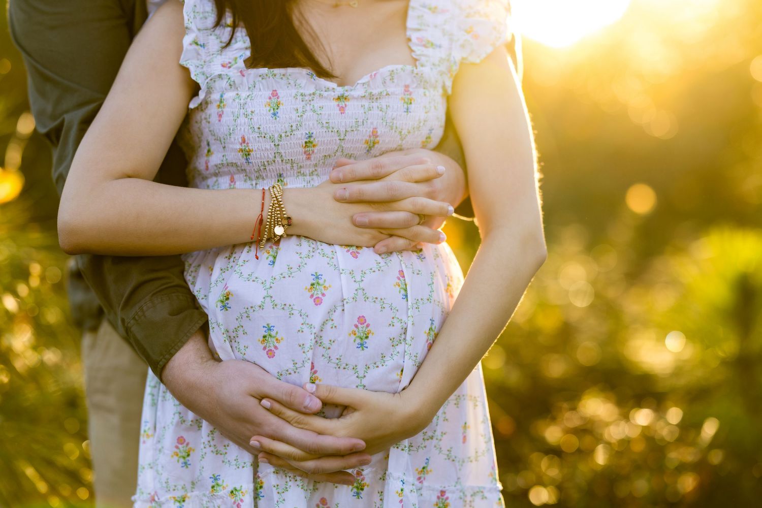 Pregnant woman in floral dress cradled by partner in golden sunset light, maternity photography outdoors.