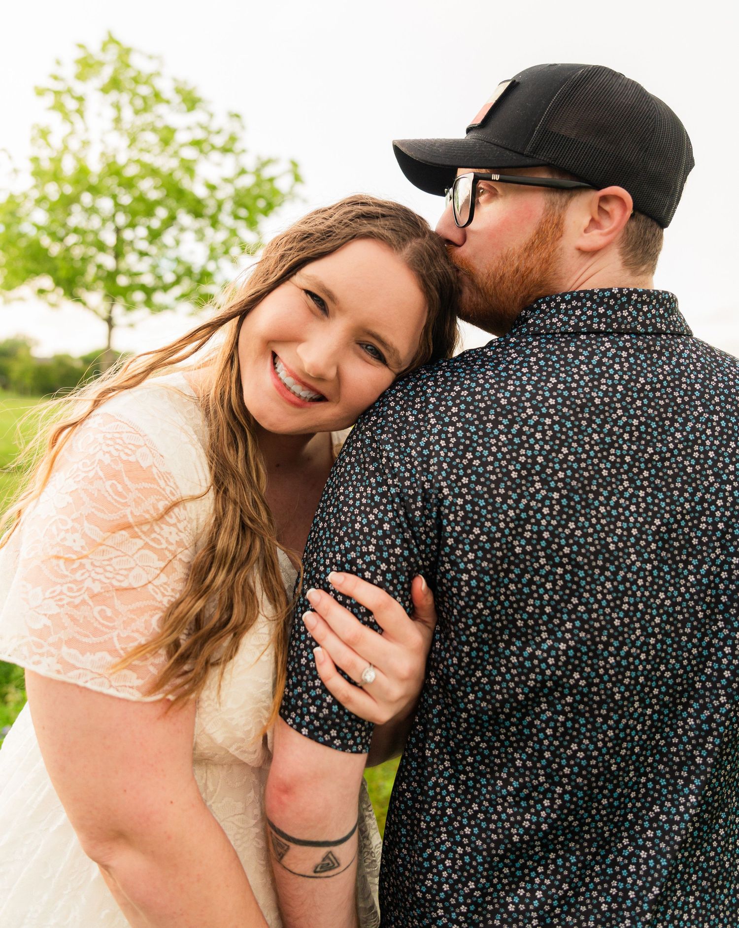 A smiling bride in a white lace dress leans into her partner who kisses her cheek outdoors.