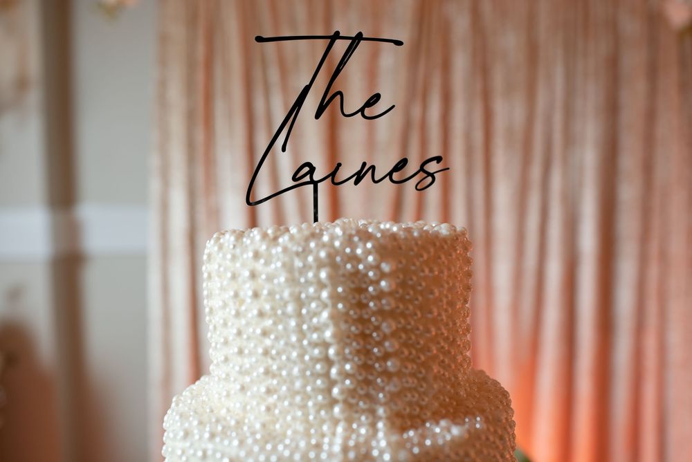 Elegant pearl-covered wedding cake with The Laines cake topper against a soft pink draped backdrop.