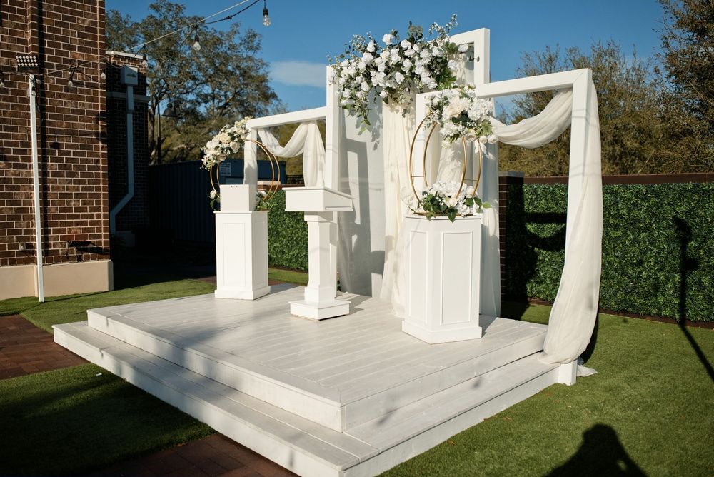 Elegant white outdoor wedding ceremony arch with floral arrangements and draped fabric on a white platform stage.