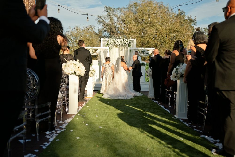 Outdoor wedding ceremony with bride and groom at floral arch, guests seated on either side of grass aisle.