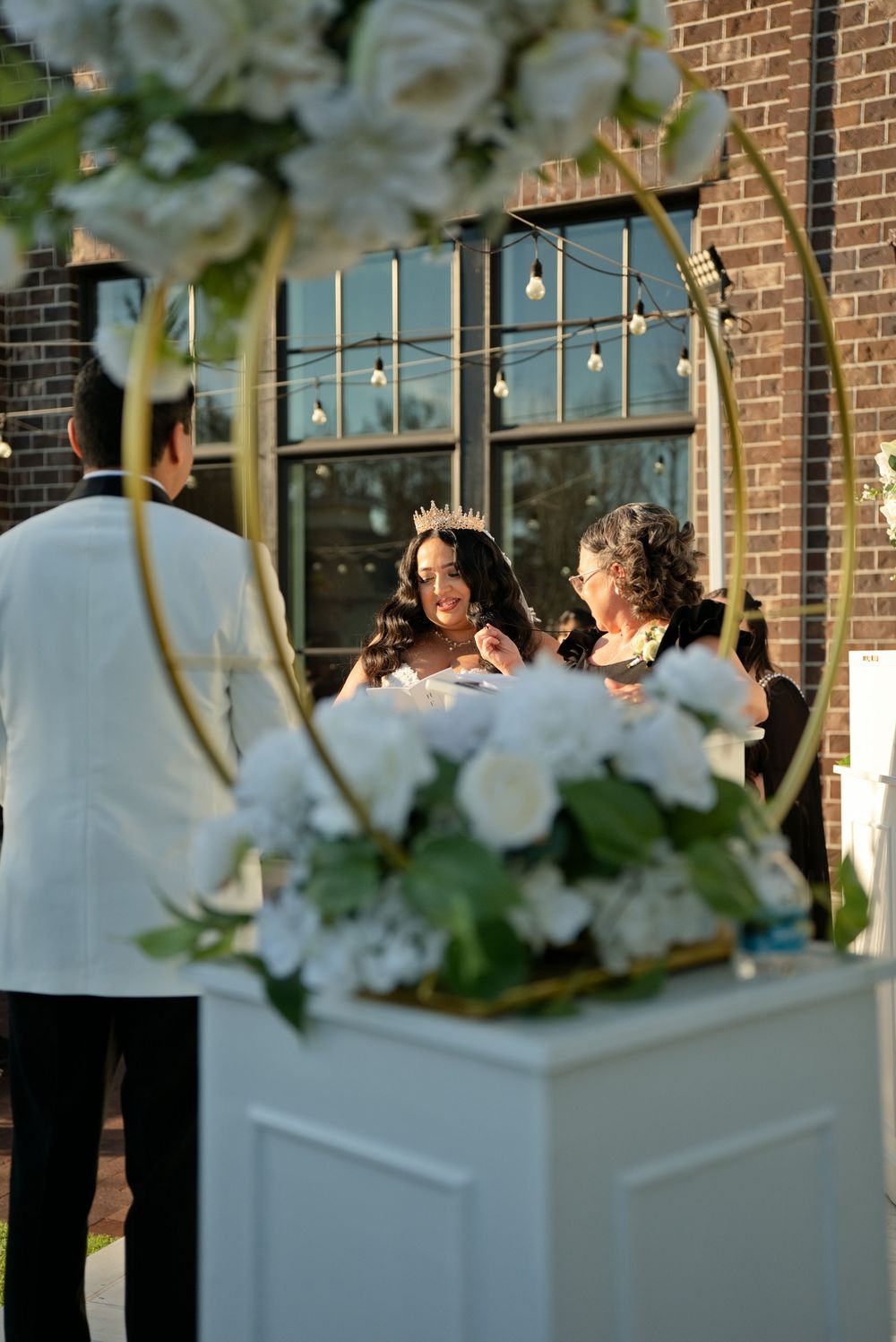 Elegant outdoor wedding ceremony with bride wearing tiara, white floral arch, and string lights against brick building backdrop.