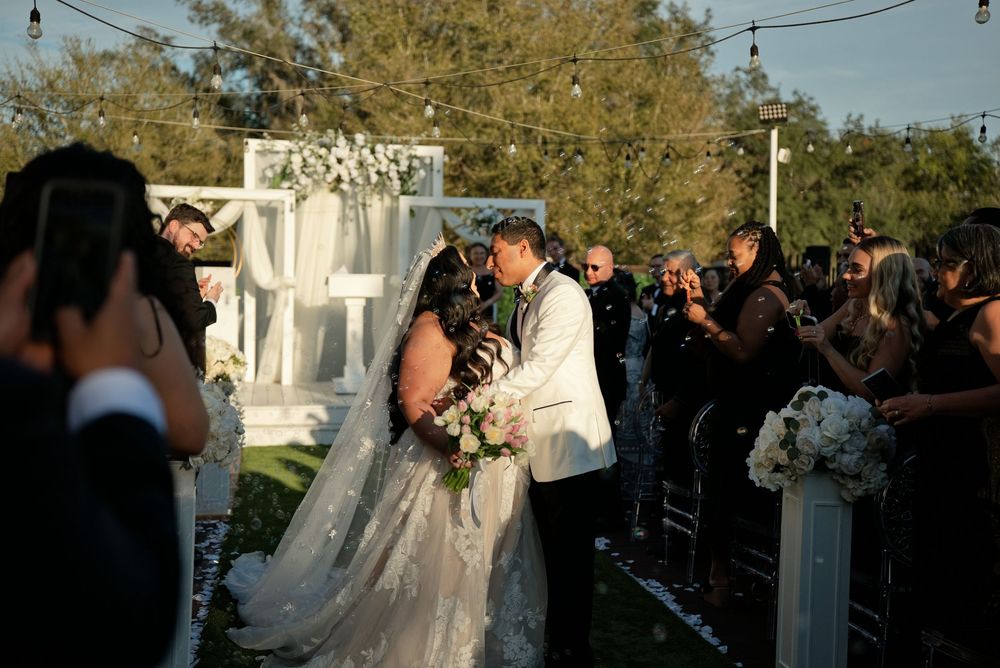 Bride and groom share first kiss at outdoor wedding ceremony with white floral arch and guests celebrating around them.