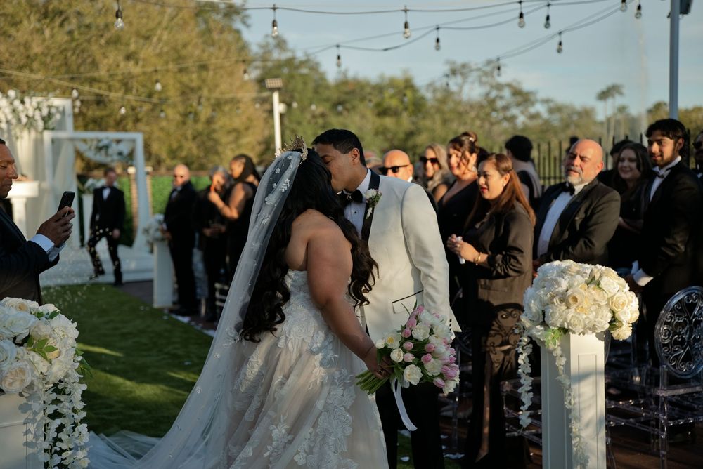 Bride and groom share a romantic kiss at an outdoor wedding ceremony surrounded by guests.