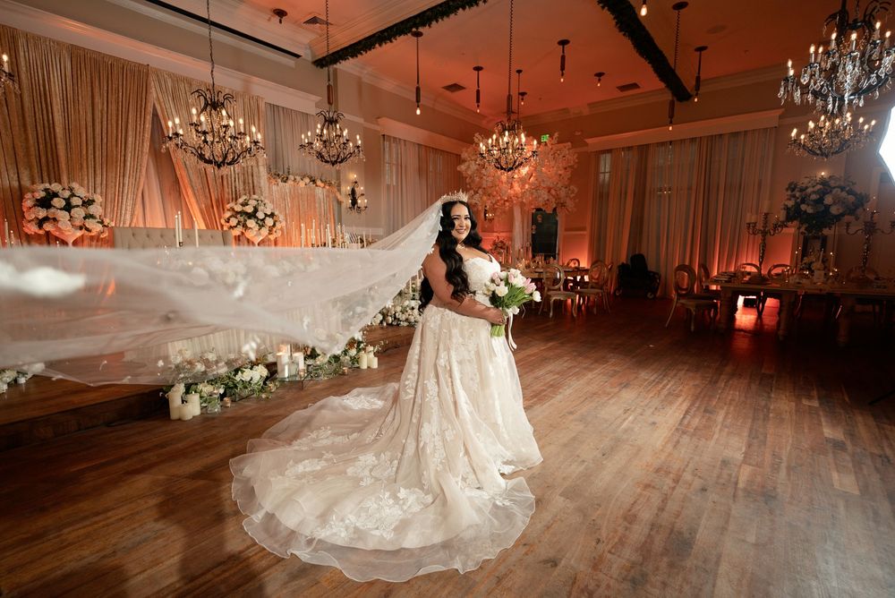 Bride in lace ballgown with flowing veil poses on wooden dance floor in elegant chandelier-lit wedding venue.