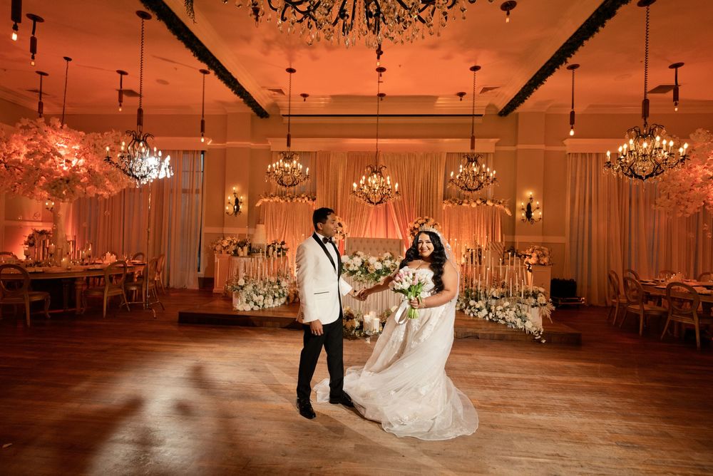 Bride and groom share first dance in elegant ballroom with chandeliers and warm pink lighting at wedding reception.