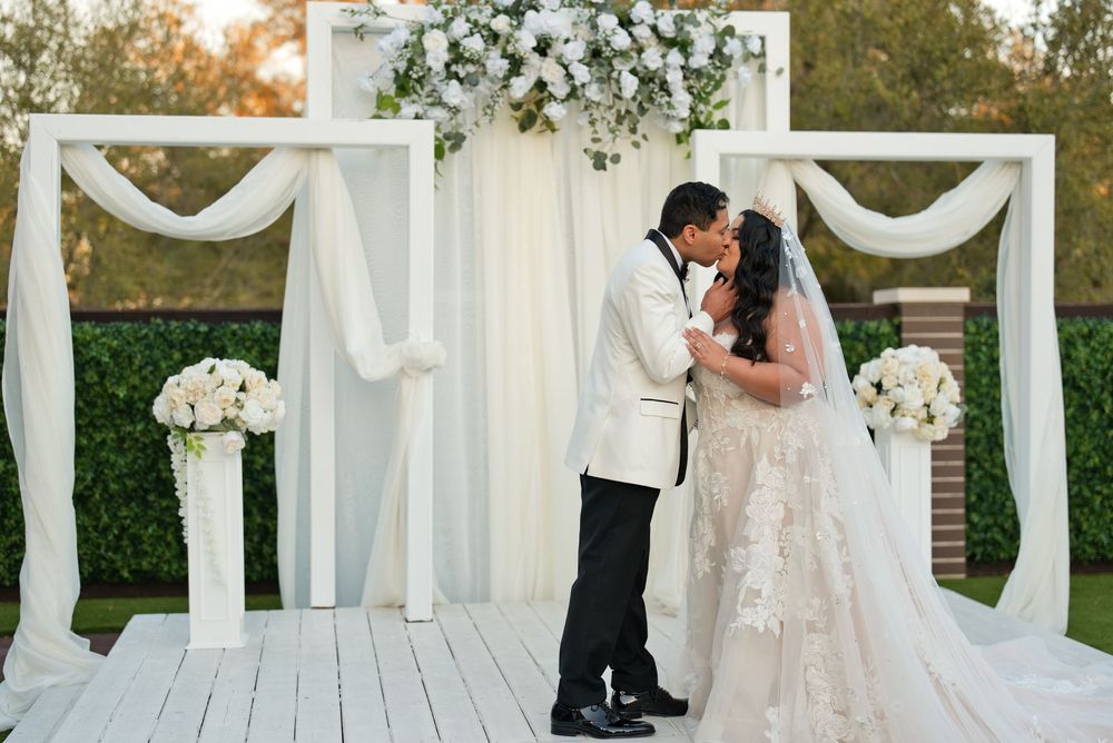 Bride and groom share first kiss at elegant outdoor wedding ceremony with white floral arch and draped fabric backdrop.