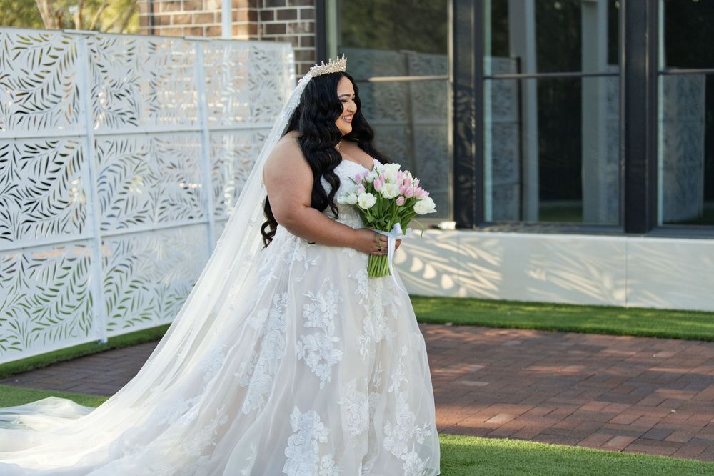 Bride in a lace ball gown with long veil holds a pink bouquet outdoors at an elegant wedding venue.