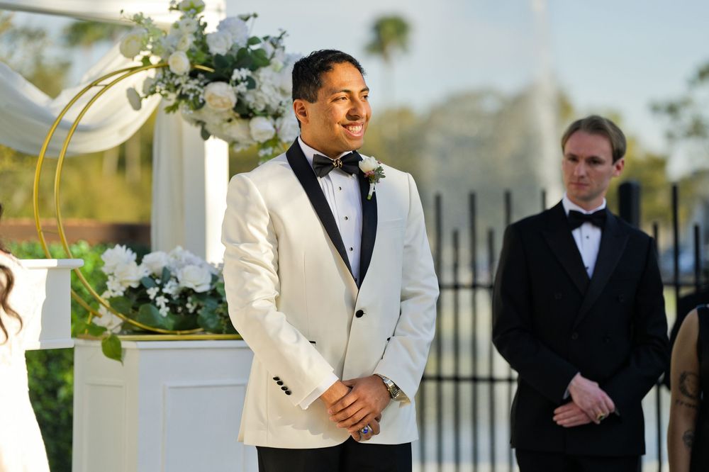 Groom in white tuxedo jacket smiling at outdoor wedding ceremony with floral arch and guest in background.