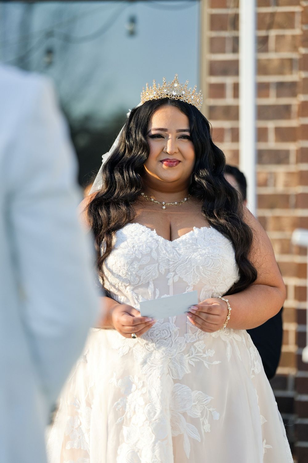 Bride in strapless lace gown and gold crown smiles while reading wedding vows outdoors near brick building.