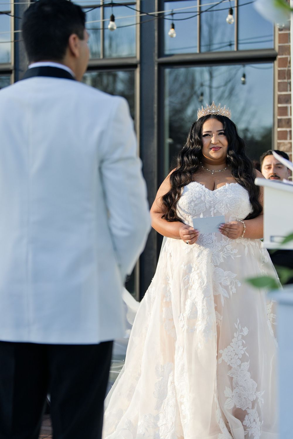 Bride in lace ballgown and tiara reads vows to groom in white tuxedo during outdoor wedding ceremony.