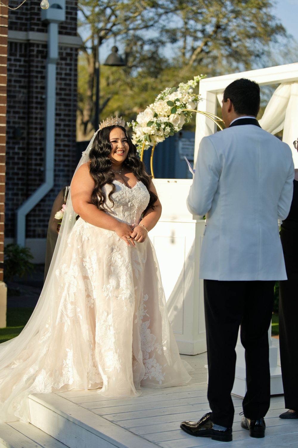 Bride in lace ball gown and crown smiles at groom in white tuxedo jacket during outdoor wedding ceremony.