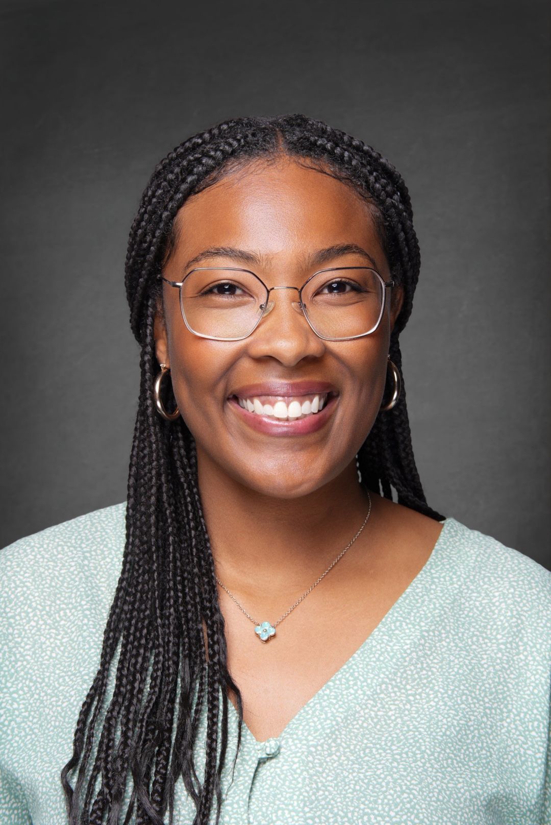 Smiling woman with long black braids, round wire glasses and hoop earrings wears a light green top in a professional headshot on gray.