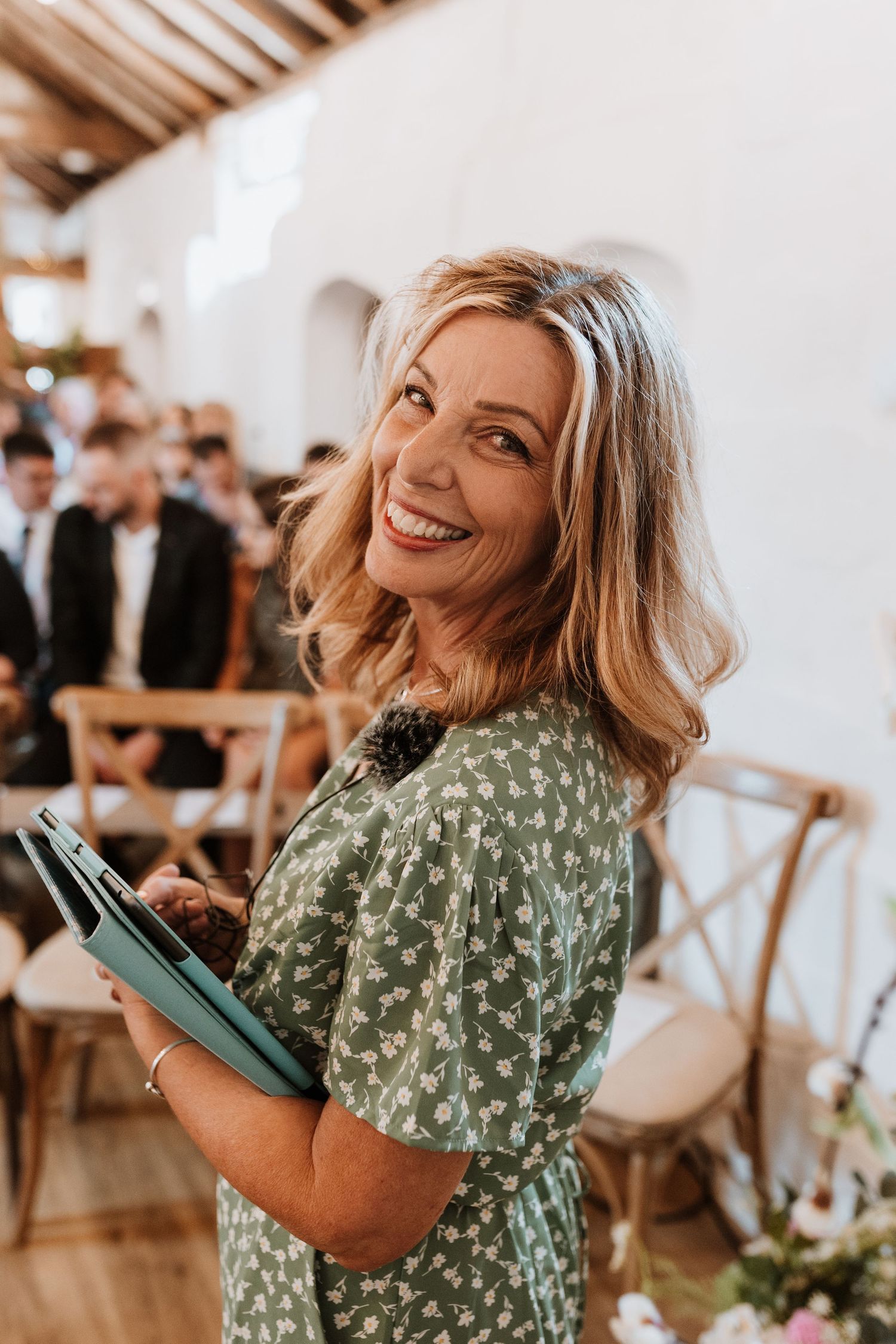 Smiling blonde woman in green floral dress holding a blue folder at an elegant wedding ceremony venue.