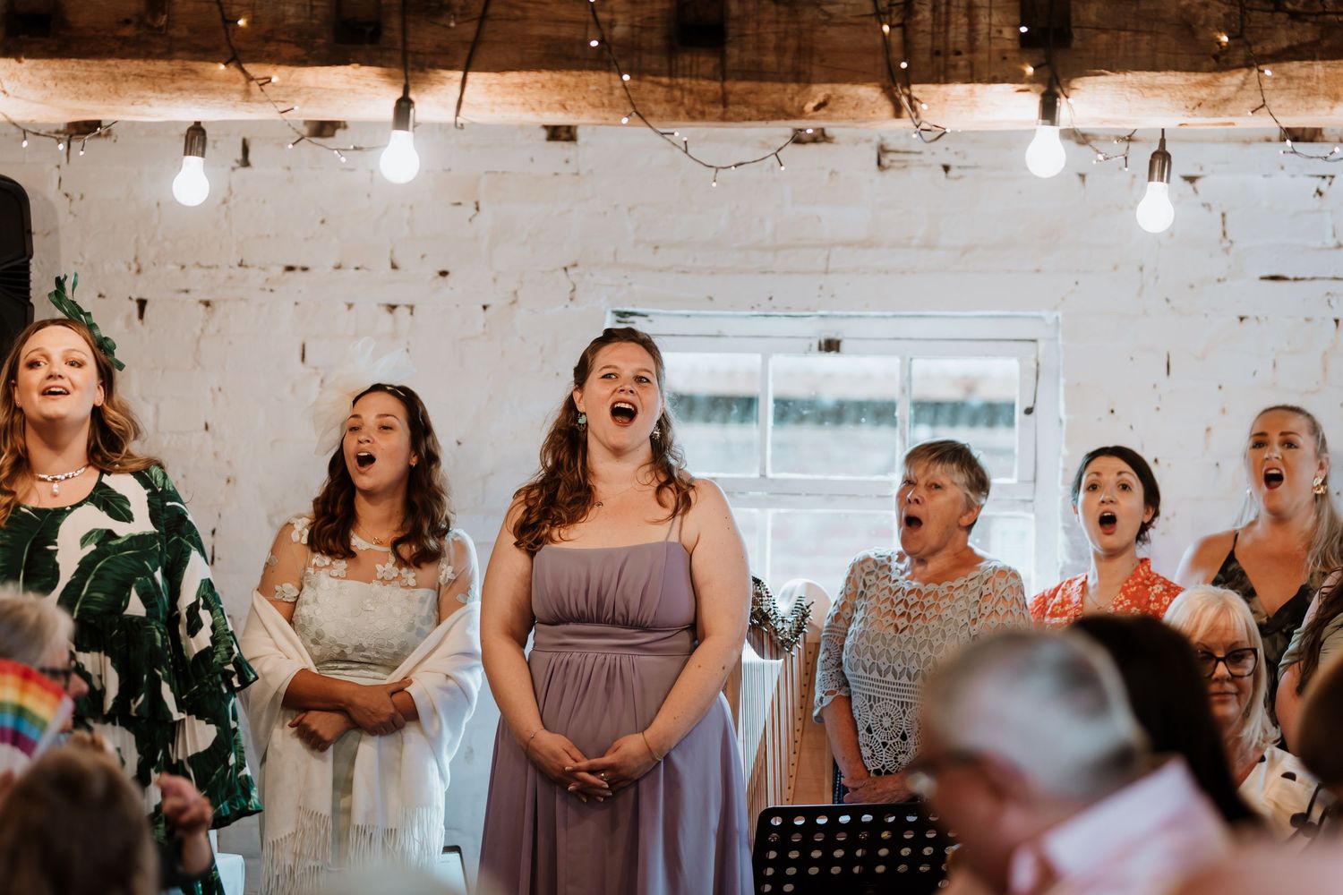 Group of women singing together at a wedding reception in a rustic white brick venue with string lights.