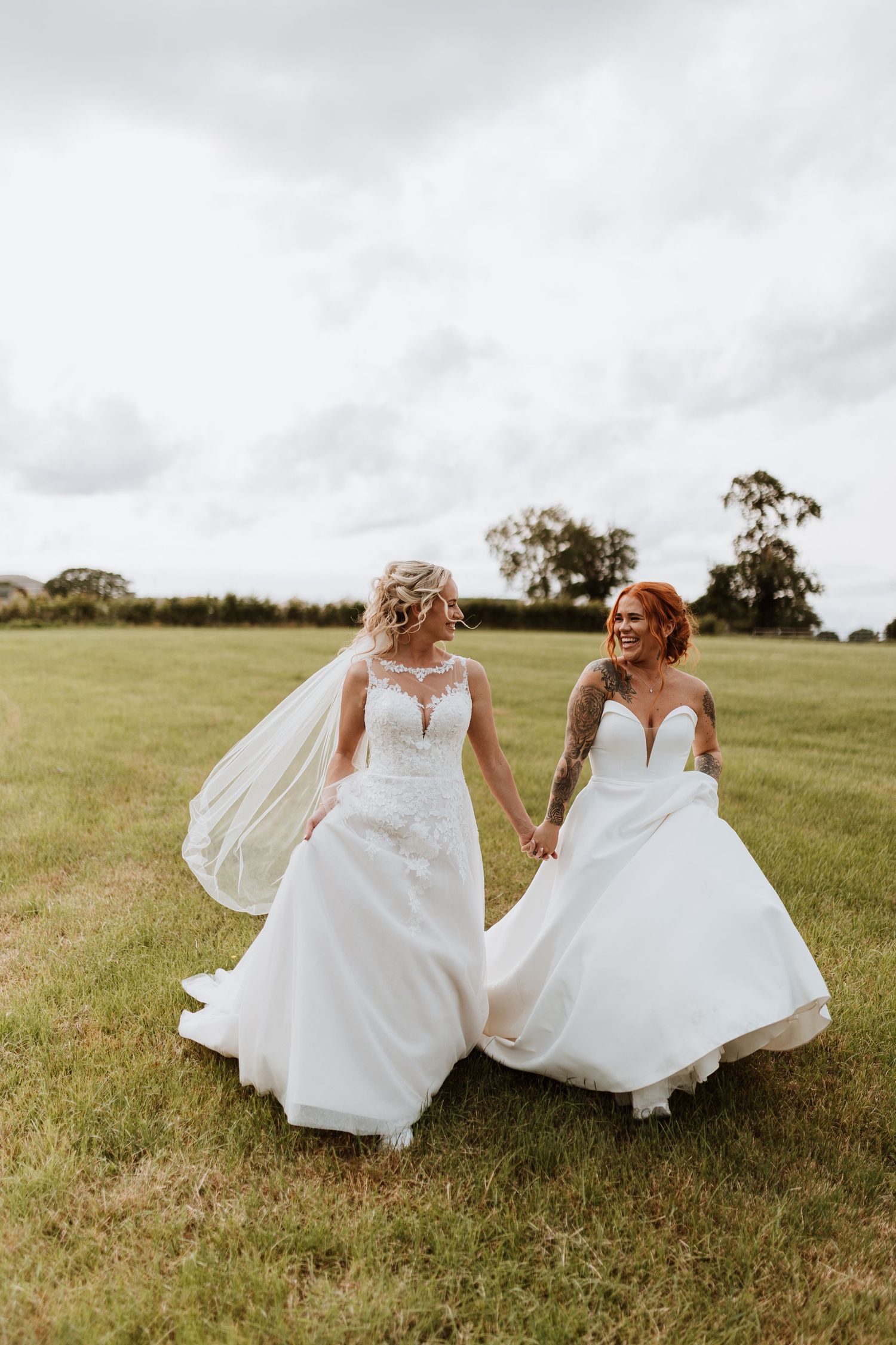 Two brides in white wedding gowns hold hands while walking through a green field on their wedding day.