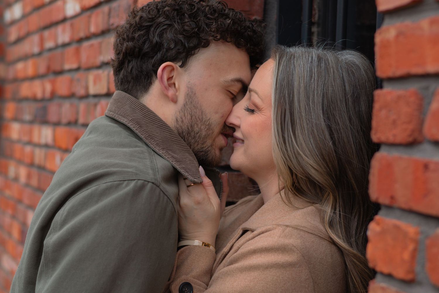 Romantic couple sharing intimate moment nearly kissing against rustic red brick wall outdoors.