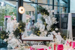 Elegant wedding reception decor with white floral arch, greenery, and pink gift bags on decorated tables.