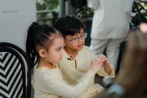 Two children in matching cream outfits sit together, with a boy in glasses holding hands with a younger girl.