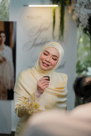 A smiling woman in a yellow hijab and outfit holds a small item at an elegant event with floral decor.