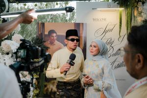 Man in traditional Malay attire speaking into microphone at elegant wedding event with floral decorations.