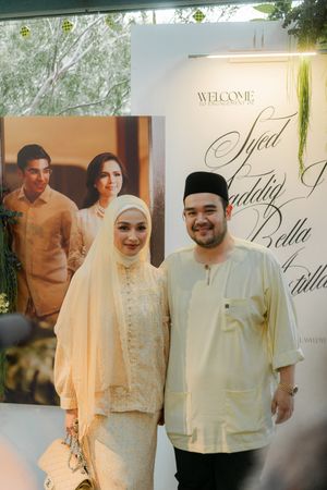 Muslim couple in cream traditional attire posing at a wedding welcome backdrop.