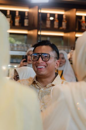 Smiling man wearing glasses and striped shirt at indoor gathering with people in background.