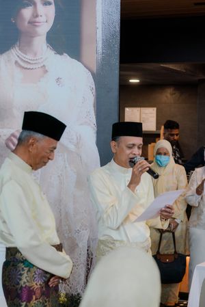 Two men in traditional Malay baju melayu and songkok caps, one speaking into a microphone at a formal ceremony.