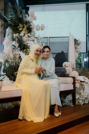 Two women in traditional Malay attire sitting together at a floral wedding ceremony decoration backdrop.
