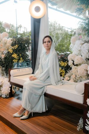 Bride in light blue modest gown and veil seated on white bench surrounded by white floral arrangements.