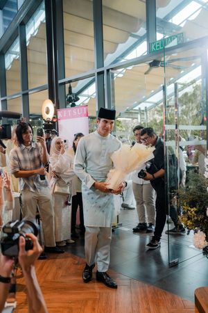 Man in traditional Malay baju melayu holding bouquet at elegant wedding event.