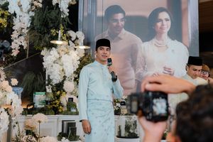 Man in light blue baju melayu giving speech at decorated wedding reception with floral arrangements.