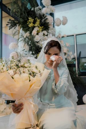 Emotional bride in light blue hijab holding yellow bouquet, wiping tears at floral decorated wedding ceremony.