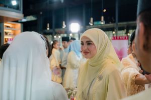 Woman in cream hijab smiling at a traditional Malay wedding celebration indoors.