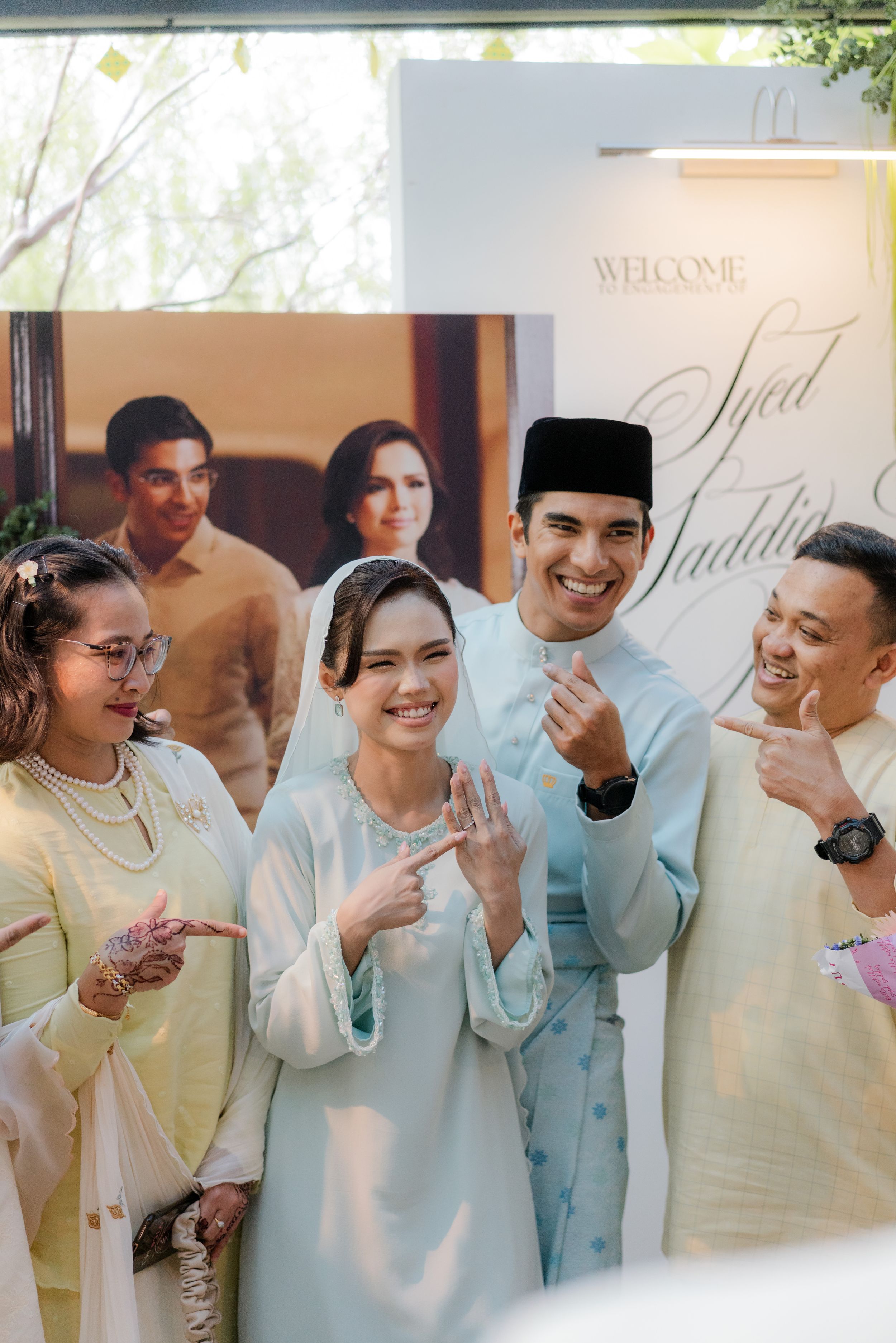 Happy Malay couple in traditional attire showing engagement ring with guests at welcome sign backdrop.